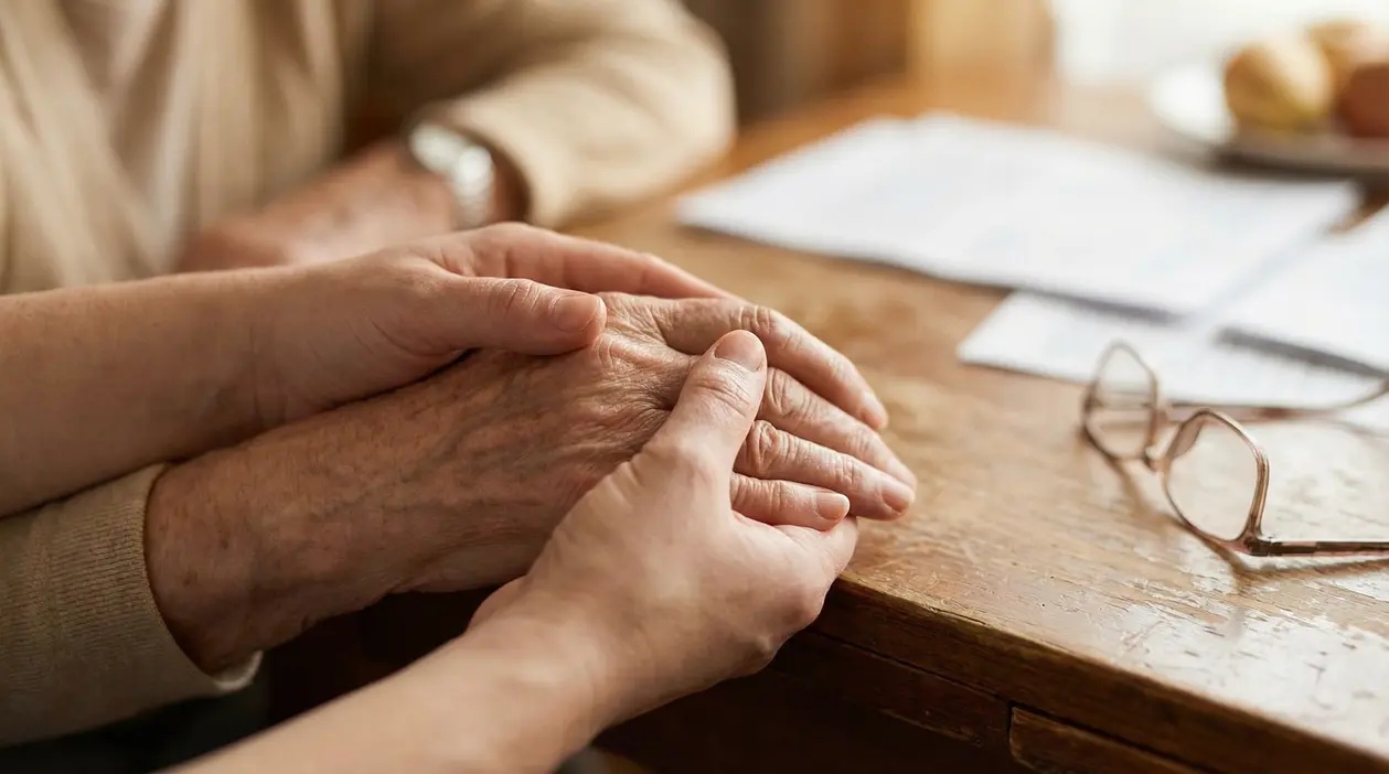 Mani di una persona che offre supporto a un anziano seduti a un tavolo di legno